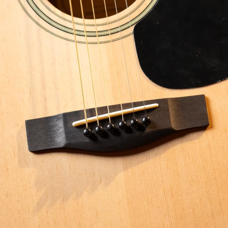 Close-up of an acoustic guitar bridge with six strings anchored by black bridge pins and a dark pickguard nearby. ขายราคาพิเศษ