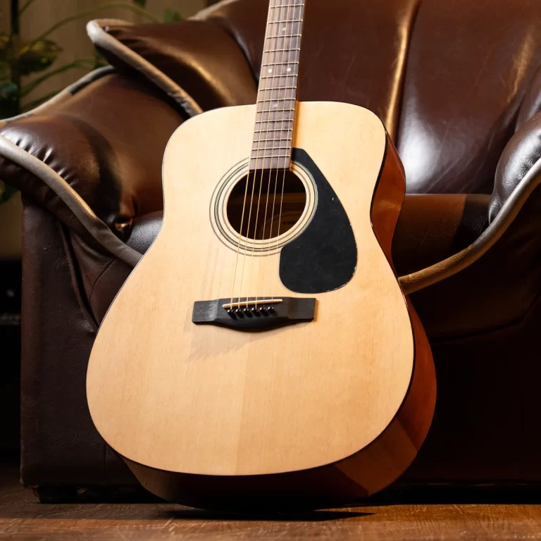 Acoustic guitar resting against a brown leather sofa, front view with soundhole, pickguard, and bridge visible. ขายราคาพิเศษ