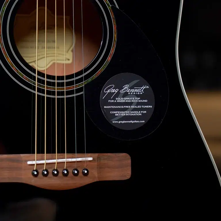Close-up of a black acoustic guitar showing the soundhole, strings, and wooden bridge; a circular label reads 'Greg Bennett Design' on the body side. ขายราคาพิเศษ