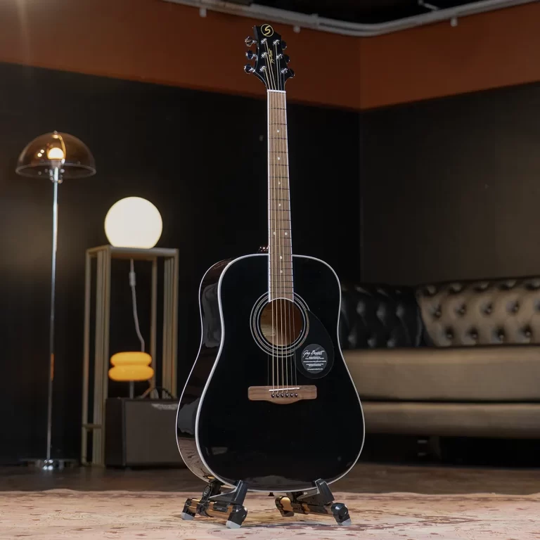 Black acoustic guitar on a stand in a stylish studio lounge with a tufted sofa and warm lighting in the background. ขายราคาพิเศษ