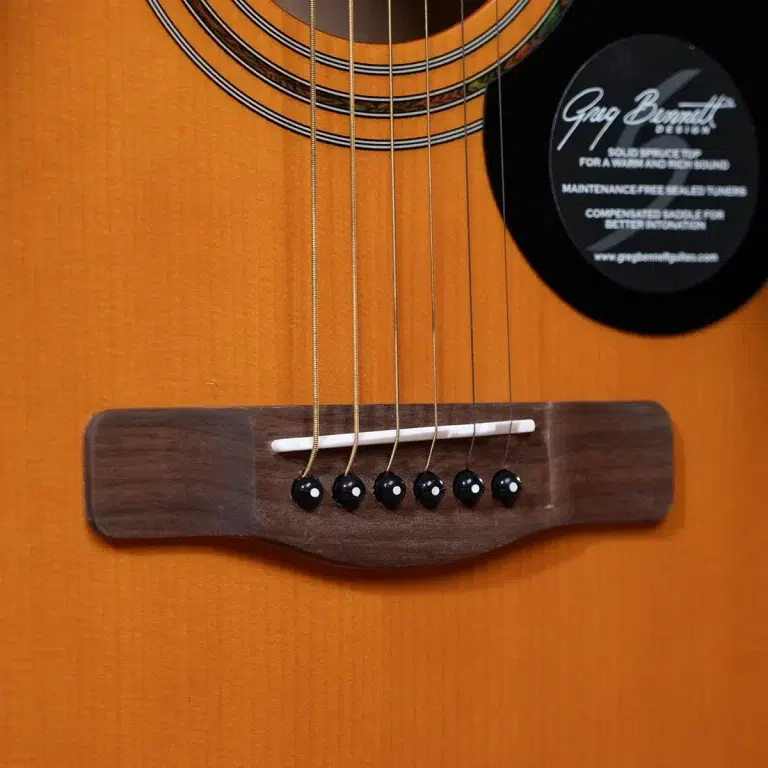 Close-up of an orange acoustic guitar showing the bridge, six strings, white saddle, and black bridge pins on a bright wood finish. ขายราคาพิเศษ