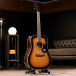 Sunburst acoustic guitar on a stand in a dim studio lounge with warm lighting and a tufted sofa in the background. ขายราคาพิเศษ