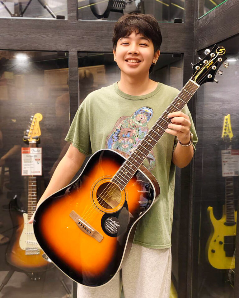 Smiling young person holding a sunburst acoustic guitar in a guitar shop, with other guitars on display in the background