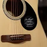 Close-up of an acoustic guitar showing the soundhole, six strings, and a black circular label on the body reading 'Greg Bennett Design'. ขายราคาพิเศษ