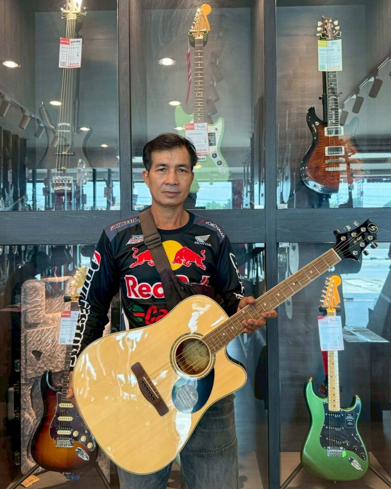 Man in a guitar shop stands holding a light-colored acoustic guitar in front of a glass display with electric guitars hanging behind him.
