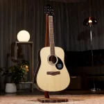 Acoustic guitar on a wooden stand in a cozy room with a warm lamp and plants in the background ลดราคาพิเศษ