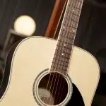 Close-up of an acoustic guitar showing the neck and fretboard with strings over the soundhole and a pale top panel under warm lighting. ขายราคาพิเศษ