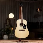 Acoustic guitar on a stand in a dimly lit room with a warm table lamp and plants in the background ขายราคาพิเศษ