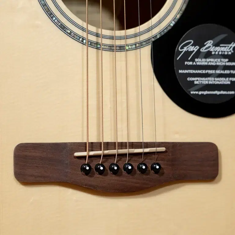 Close-up of an acoustic guitar bridge with six black pins and a white saddle on a light wood body, strings running toward the soundhole left. ขายราคาพิเศษ