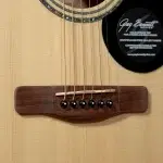 Close-up of an acoustic guitar bridge with six strings anchored to the bridge pins on a light wood top, with a circular sticker on the right side of the soundboard. ขายราคาพิเศษ