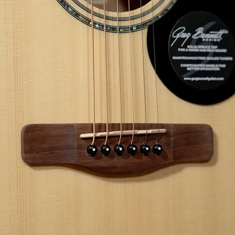 Close-up of an acoustic guitar bridge with six strings anchored to the bridge pins on a light wood top, with a circular sticker on the right side of the soundboard. ขายราคาพิเศษ