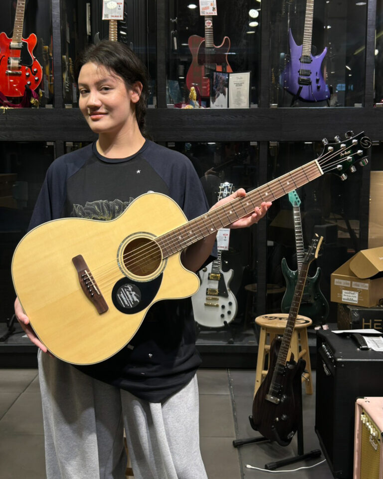 Young woman in a guitar shop holds an acoustic guitar, with a wall of electric guitars on display behind her.