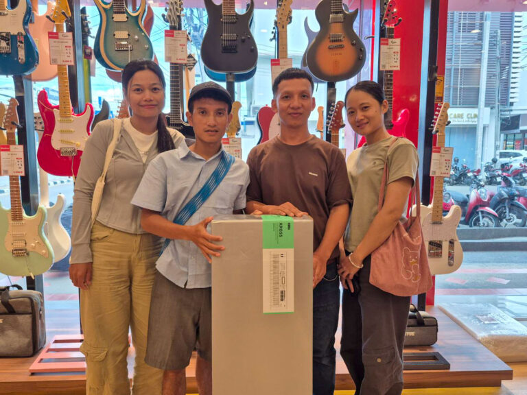 Four people smile and pose in a guitar store, standing around a large boxed item with guitars on display behind them.