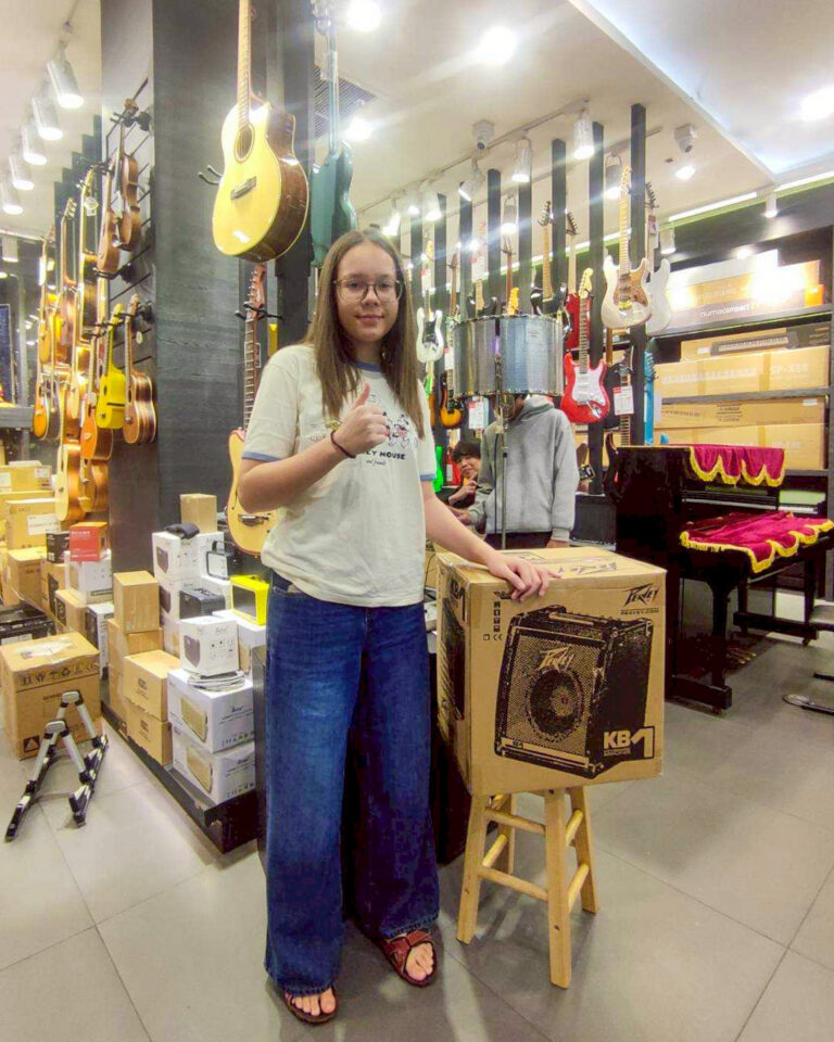 Young woman in a guitar shop giving a thumbs-up beside a boxed Peavey KB1 amplifier on a stool, with guitars hanging on racks behind her.