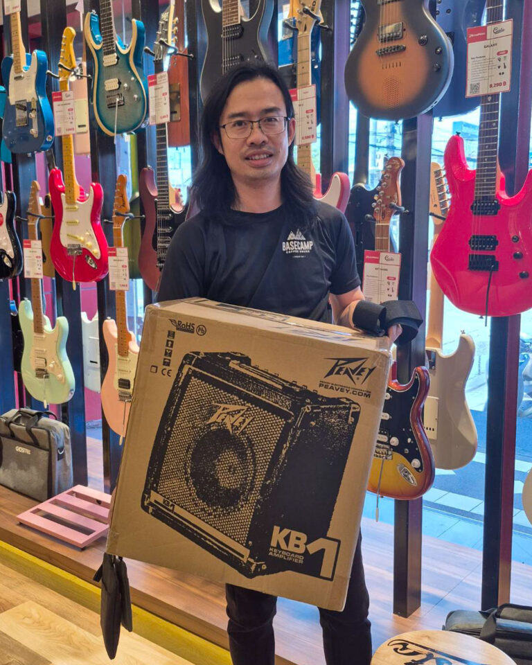 Man in a black BASECAMP shirt holds a large Peavey KB1 keyboard amplifier box in a guitar shop.