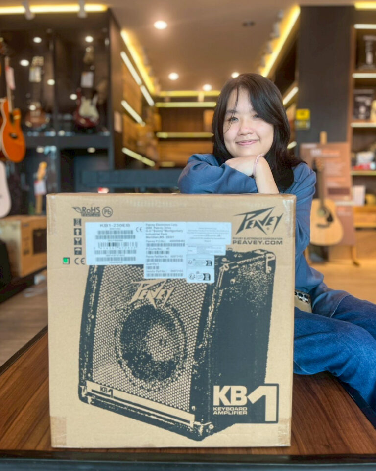 Woman in a music store smiles while resting her chin on a large box labeled KB1 keyboard amplifier