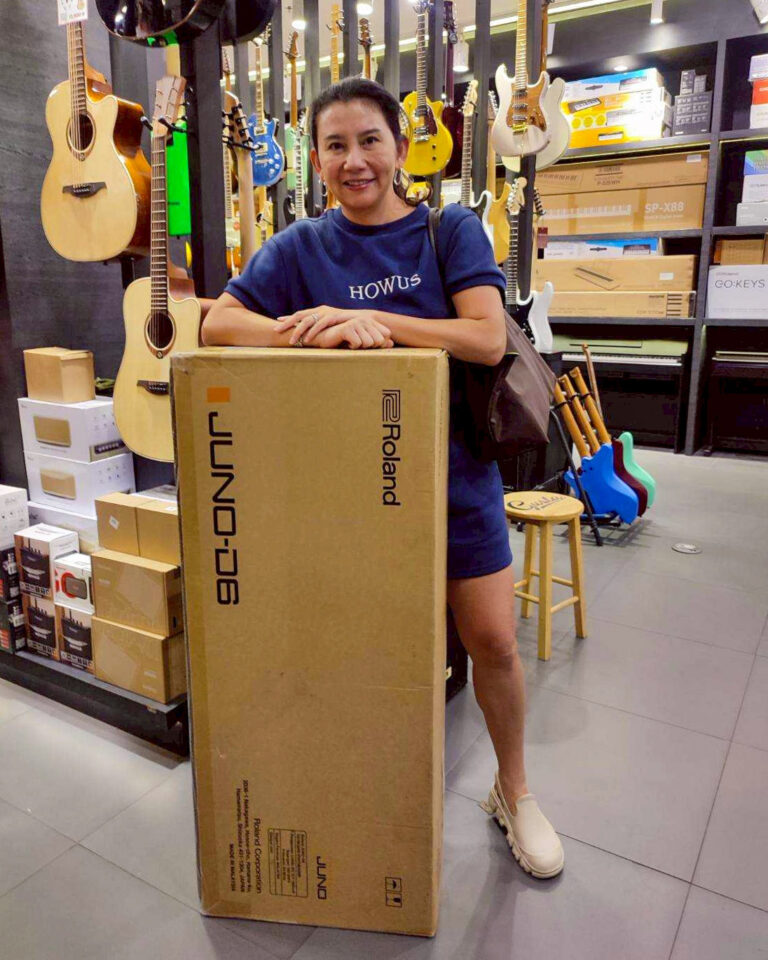 Woman in a guitar shop leaning on a large cardboard box labeled Roland JUNO/keyboard, surrounded by guitars and gear on shelves.