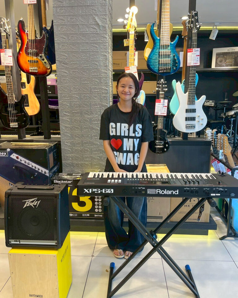 Girl standing at a Roland XPS-60 keyboard in a music store, with colorful guitars on display behind her.
