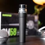 Black Shure SM58 microphone standing upright on a table with its capsule and metal grille visible, blurred box in the background. ขายราคาพิเศษ