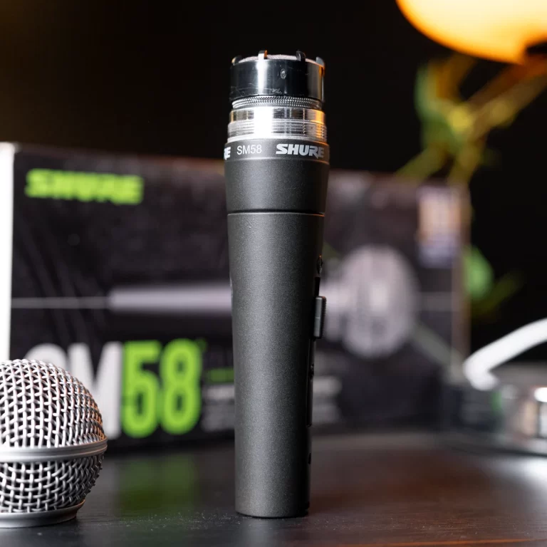 Black Shure SM58 microphone standing upright on a table with its capsule and metal grille visible, blurred box in the background. ขายราคาพิเศษ