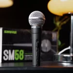 Shure SM58 microphone standing upright on a dark desk with its metal grille visible and a blurred box in the background. ลดราคาพิเศษ