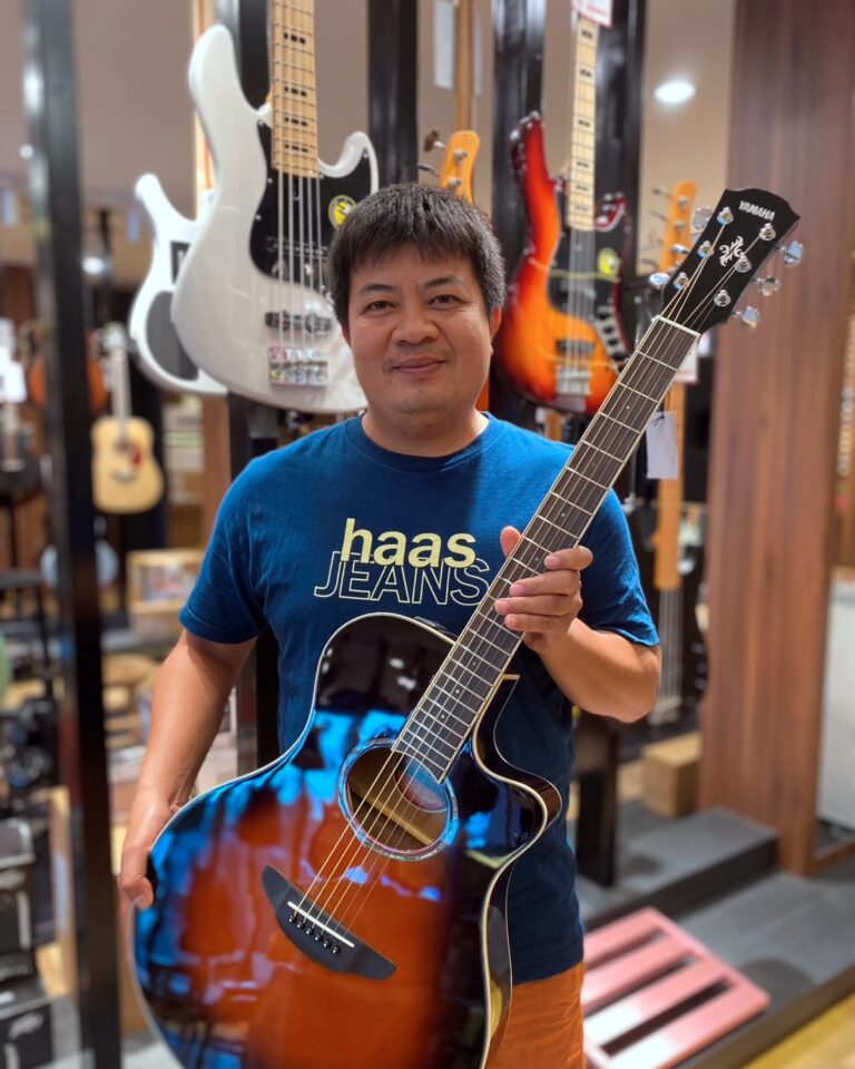 Man in a guitar shop holding a sunburst acoustic guitar, smiling at the camera.
