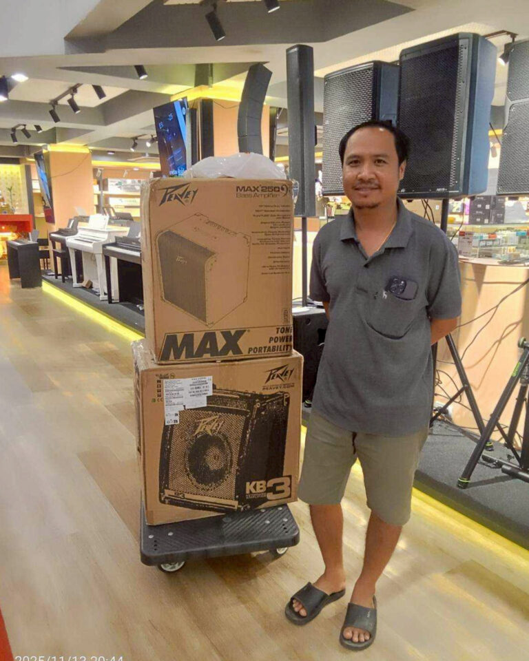 Man in gray polo and shorts stands next to a cart with two boxed Peavey MAX 250 amplifiers in a music store.