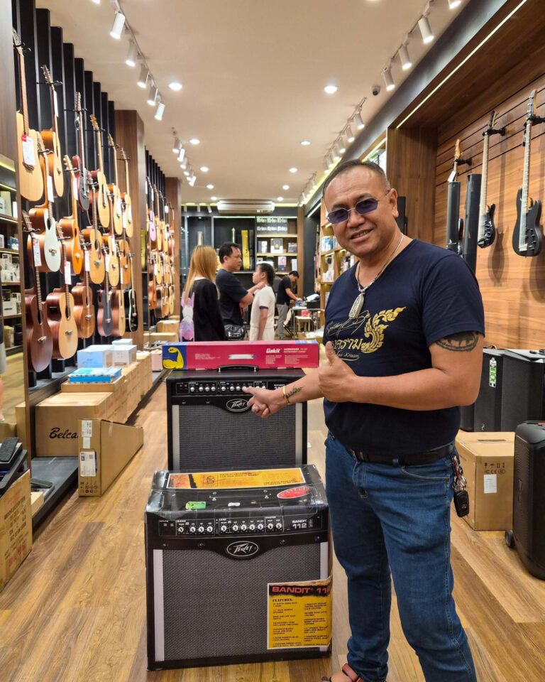 Smiling man in a guitar shop giving a thumbs-up beside a stack of Peavey Bandit amplifiers and wall displays of guitars.