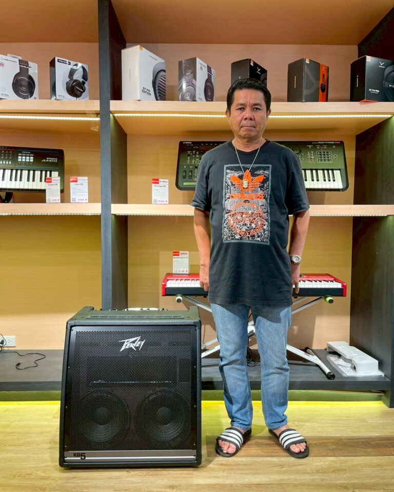 Man in a black Adidas shirt and jeans standing in a music store between shelves of keyboards and boxed headphones, with a black speaker/amp (Peavey KB5) on the floor in front of him