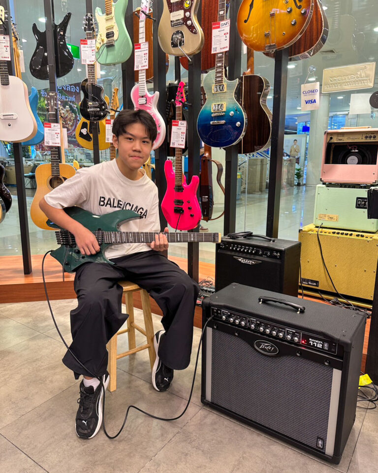 Young person seated on a stool playing a green electric guitar in a guitar shop, with amps and colorful guitars on display behind them.
