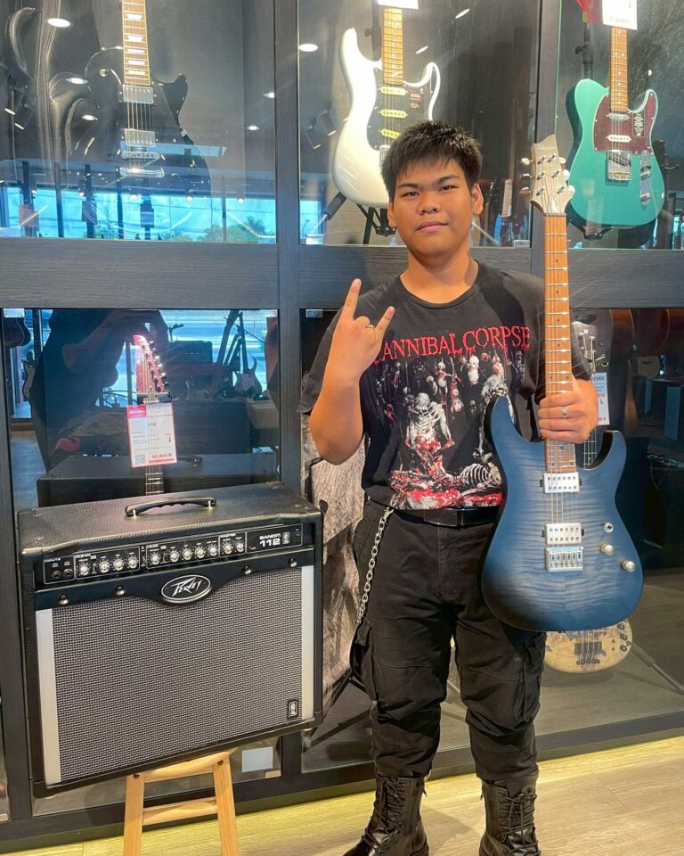 Young man in a black Cannibal Corpse T‑shirt posing with a blue electric guitar in a music store, with amplifiers and other guitars on display behind him.