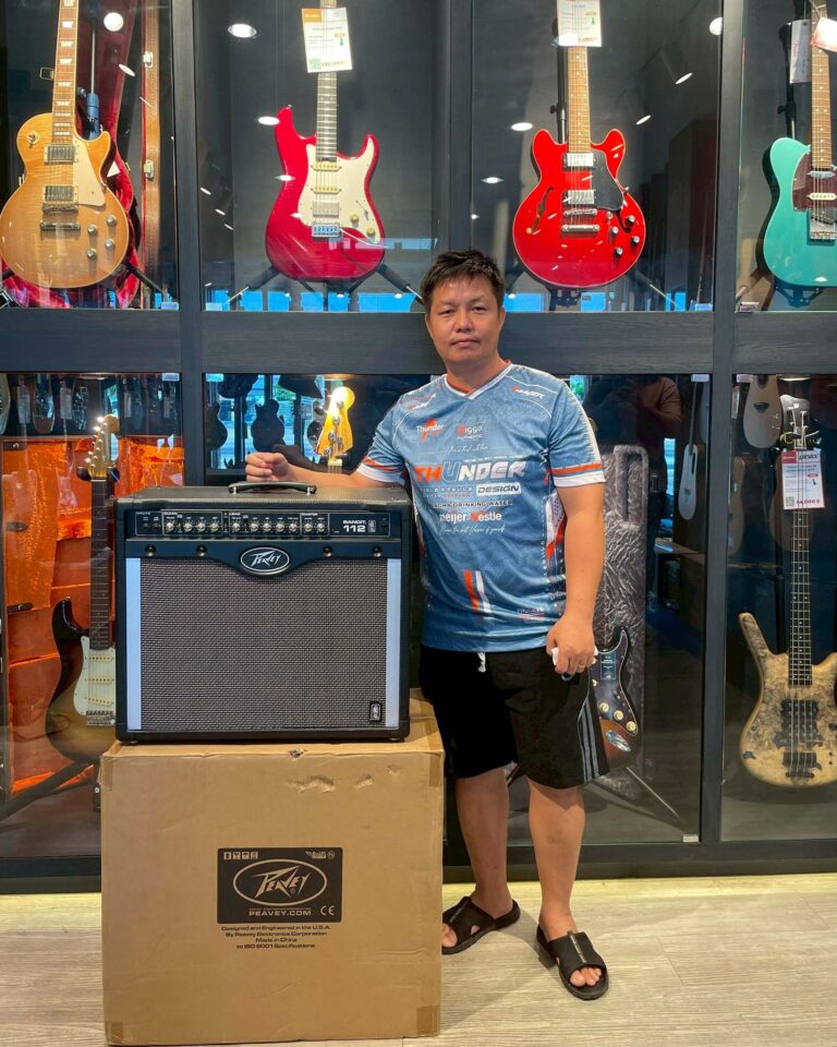 Asian man in a blue shirt stands beside a Peavey amplifier in front of a guitar display inside a music shop.