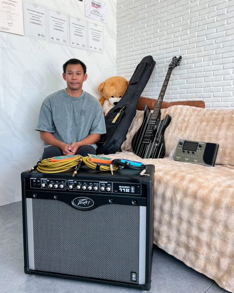 Young man sits on a couch with a striped amp, guitar, and cables in a music studio.