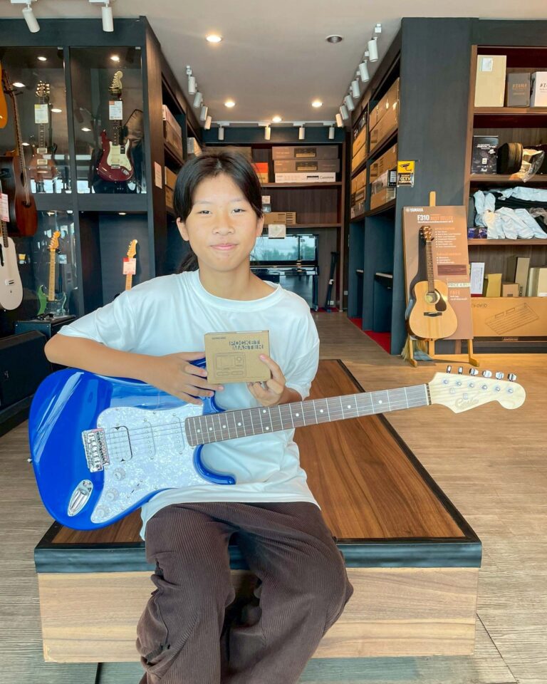 Girl in a guitar shop sits with a blue electric guitar on her lap, holding a small box and smiling at the camera.