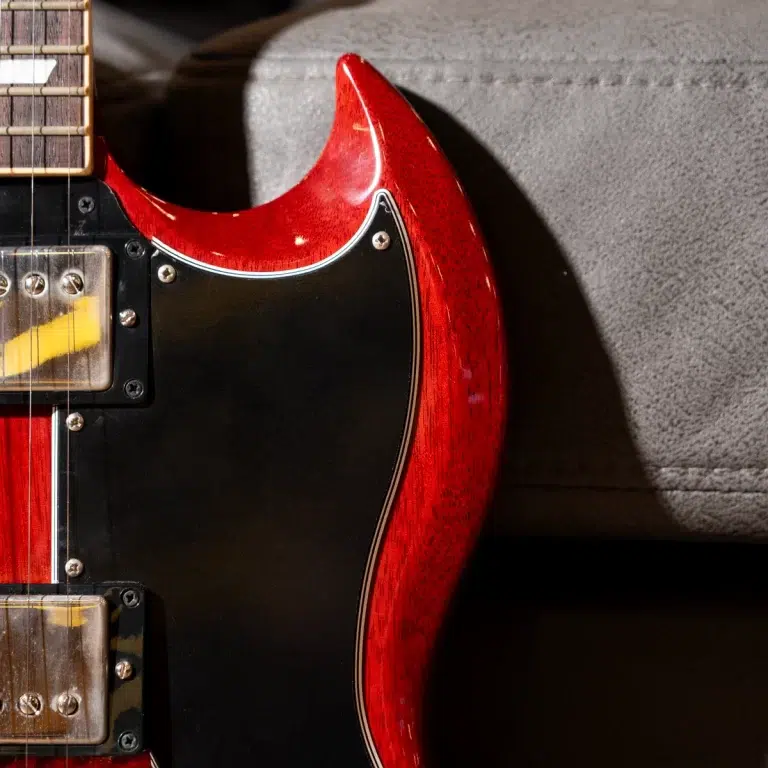 Close-up of a red electric guitar body with a black pickguard and chrome screws resting against a grey couch. ขายราคาพิเศษ