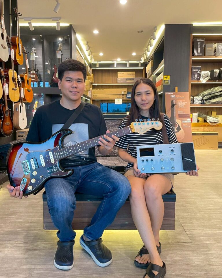 Two people pose with musical gear in a guitar shop, man holding a sunburst electric guitar and woman with a small audio mixer/synth beside her on a stand behind them.