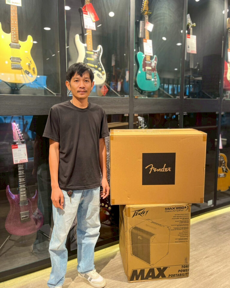 Man standing in a guitar shop next to a large Fender delivery box and a Peavey amplifier box on the floor.