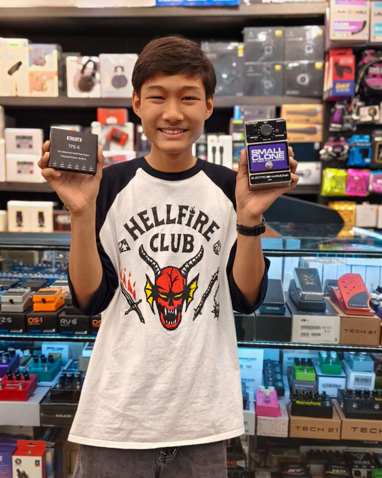 Smiling boy in a tech store holds a Small Clone guitar effects pedal in one hand and a TPS‑5 power supply box in the other.