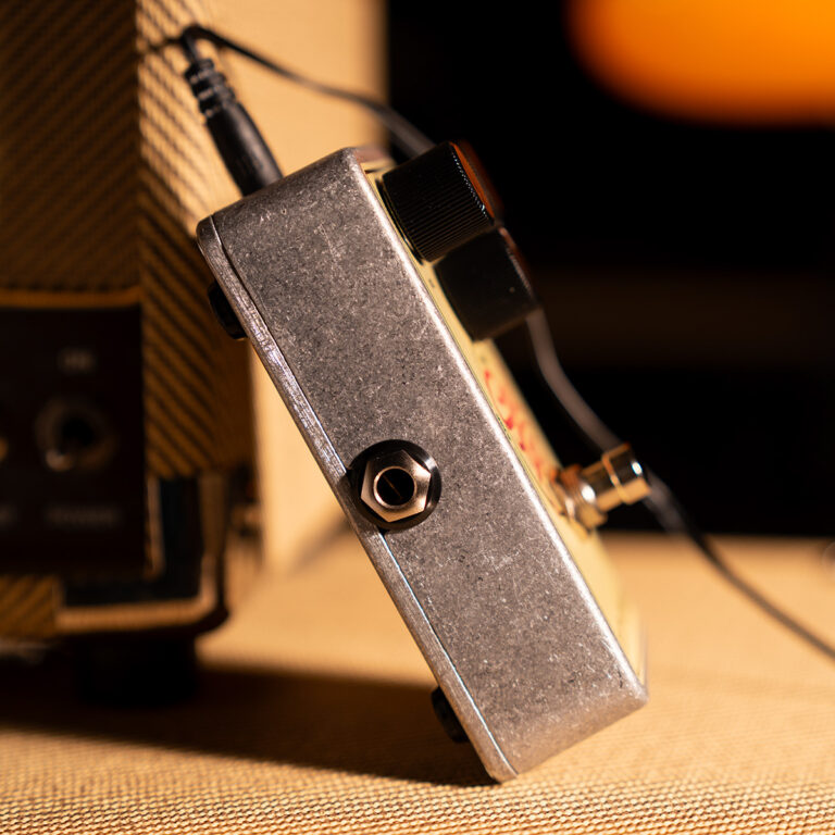 Close-up of a metal guitar effects pedal with input jack and cable plugged in, resting on a textured surface. ขายราคาพิเศษ