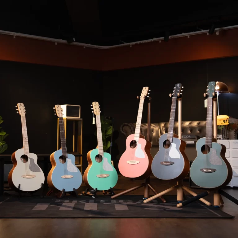 Six pastel-colored acoustic guitars on stands in a dim, warm-lit showroom/studio setting. ขายราคาพิเศษ