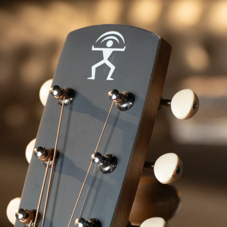Close-up of a guitar headstock with six metal tuning pegs and white plastic knobs, featuring a white stick-figure logo lifting a semicircular shape above its head on a gray headstock ขายราคาพิเศษ