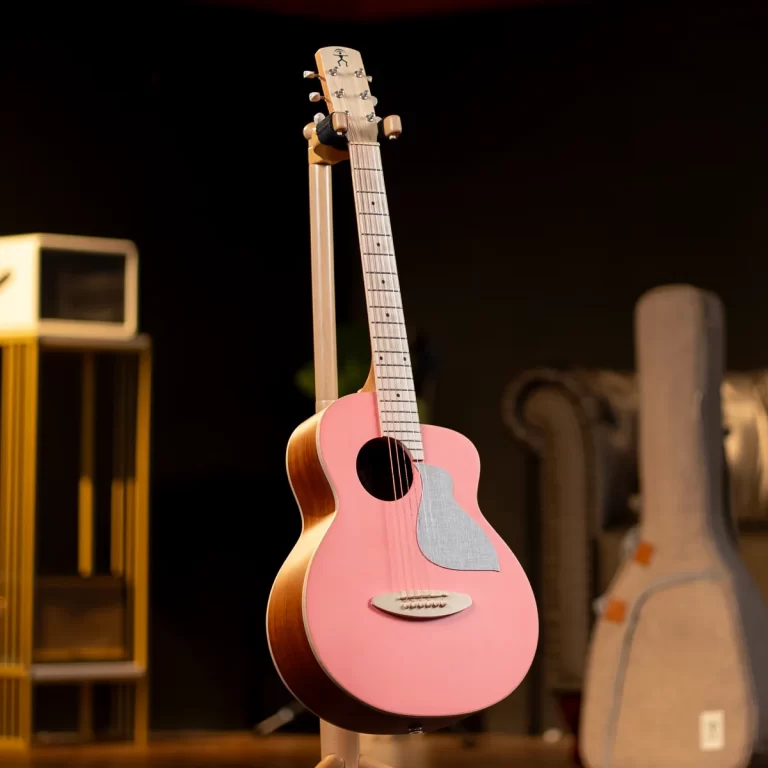 Pink acoustic guitar on a stand in a dimly lit music studio, with other guitar cases visible in the background. ขายราคาพิเศษ