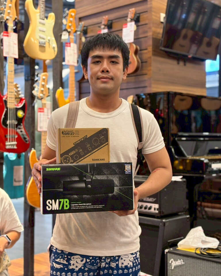 Man standing in a music store, holding a Shure SM7B microphone box and another boxed audio device in front of guitars on display behind him.