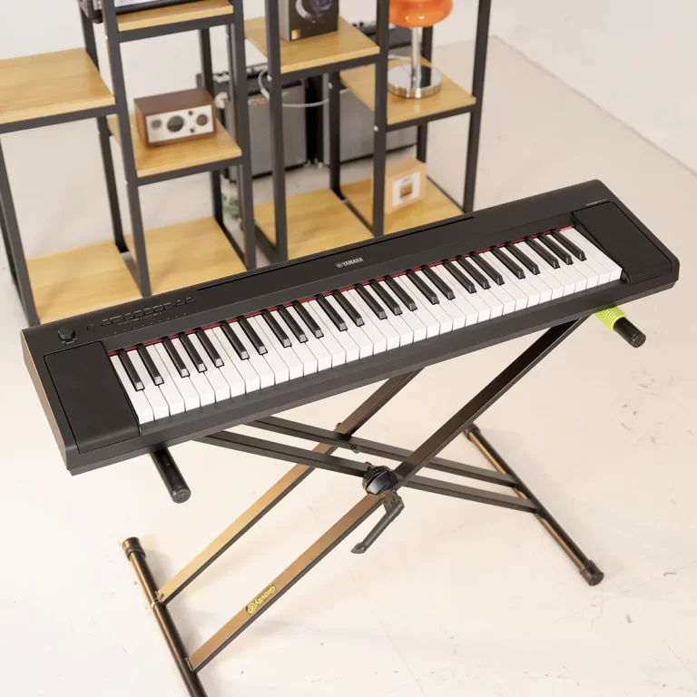 Yamaha digital keyboard on a black folding stand in a bright studio with wooden shelves in the background. ขายราคาพิเศษ