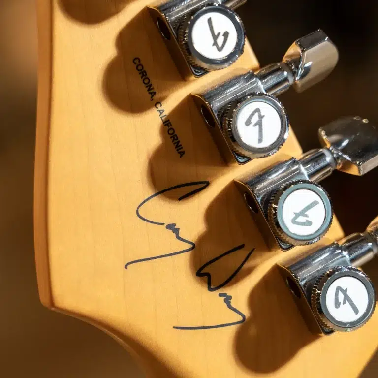 Close-up of a guitar headstock with chrome tuning pegs, circular logo on each tuner, and a handwritten signature on the wood near 'Corona, California'. ขายราคาพิเศษ