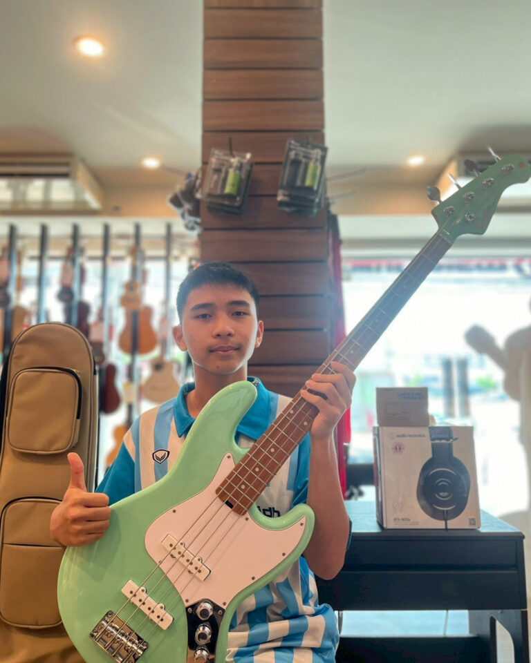 Young man in a blue striped shirt holding a mint green bass guitar in a music store, giving a thumbs up.