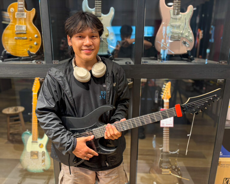 Young man in a black jacket posing with a black electric guitar in a guitar shop, with guitar displays behind him in glass cases.