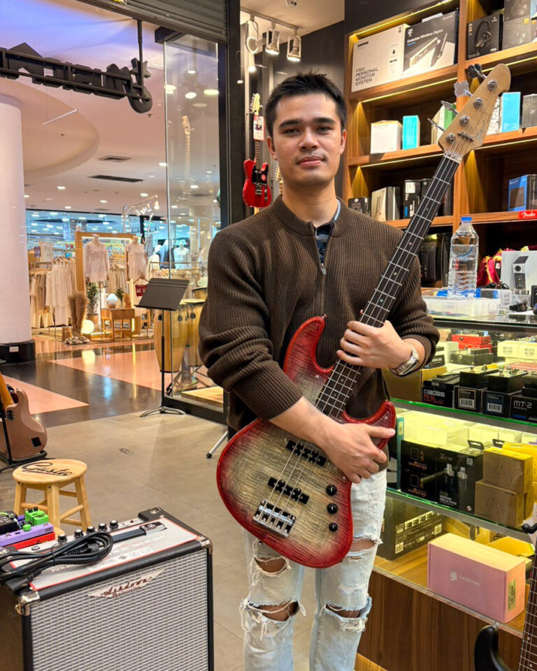 Young man in a brown sweater holding a red sunburst bass guitar in a music store.