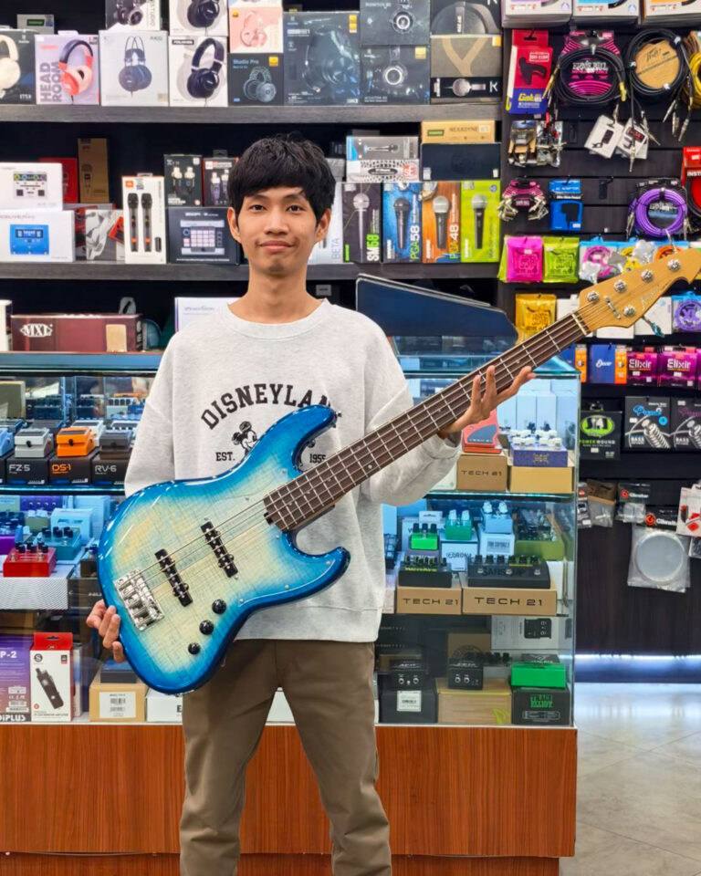 Young man in a gray sweatshirt holding a blue electric bass guitar in a music store filled with accessories and gear boxes behind him.