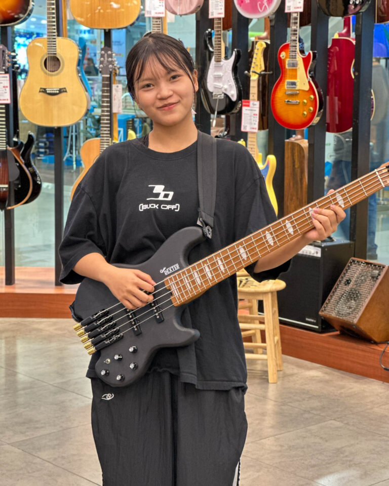 Young woman in a black oversized shirt holding a dark bass guitar in a guitar shop, with colorful guitars hanging in the background.